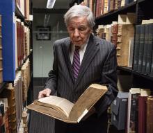 Professor Helmholz, in a dark pinstripe suit and tie, holds an old book. Shelves of rare and old books surround him.