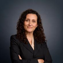 Professional headshot of Sandra Frantzen wearing a dark suit and beige blouse. Her hands are folded across her body.