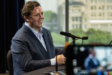 Will Baude wearing a suit is speaking into a microphone while sitting at a desk