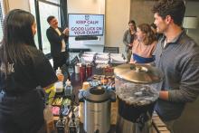 Students gathered around at table with coffee with someone pointing at a screen that says "Keep Calm and Good Luck on Your Exams".