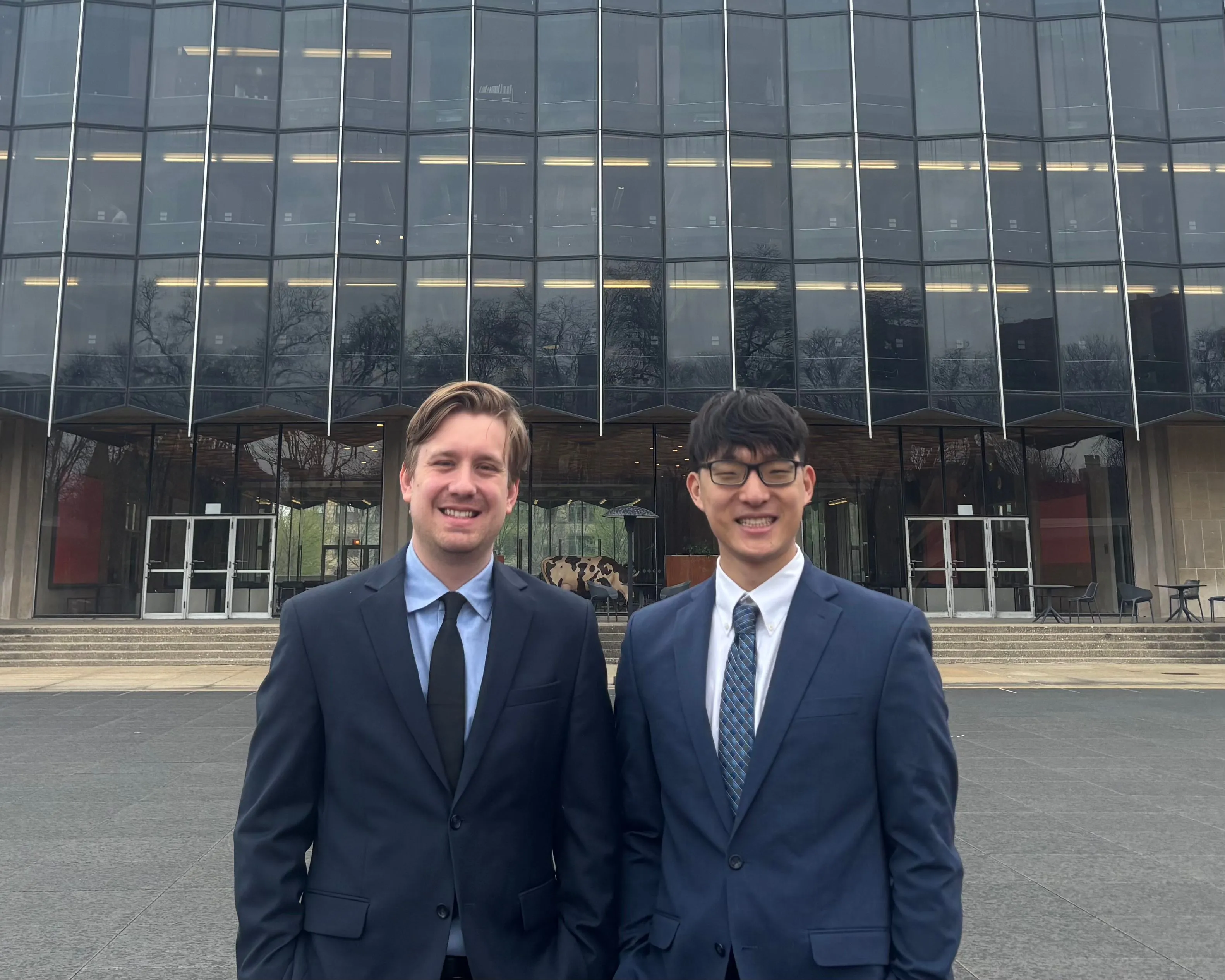 Two men wearing suits standing in front of a large glass building.