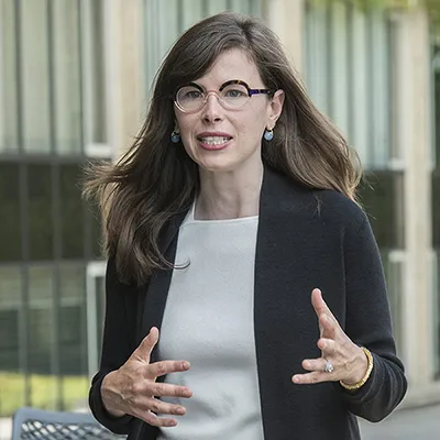 Bridget Fahey stands outside, gesturing. She wears a neutral top, and a dark sweater.