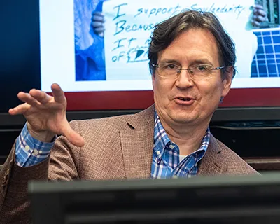 Mark Templeton makes a point with a presentation behind him and computer in front of him. He wears a blue open-collared shirt and brown sports jacket.