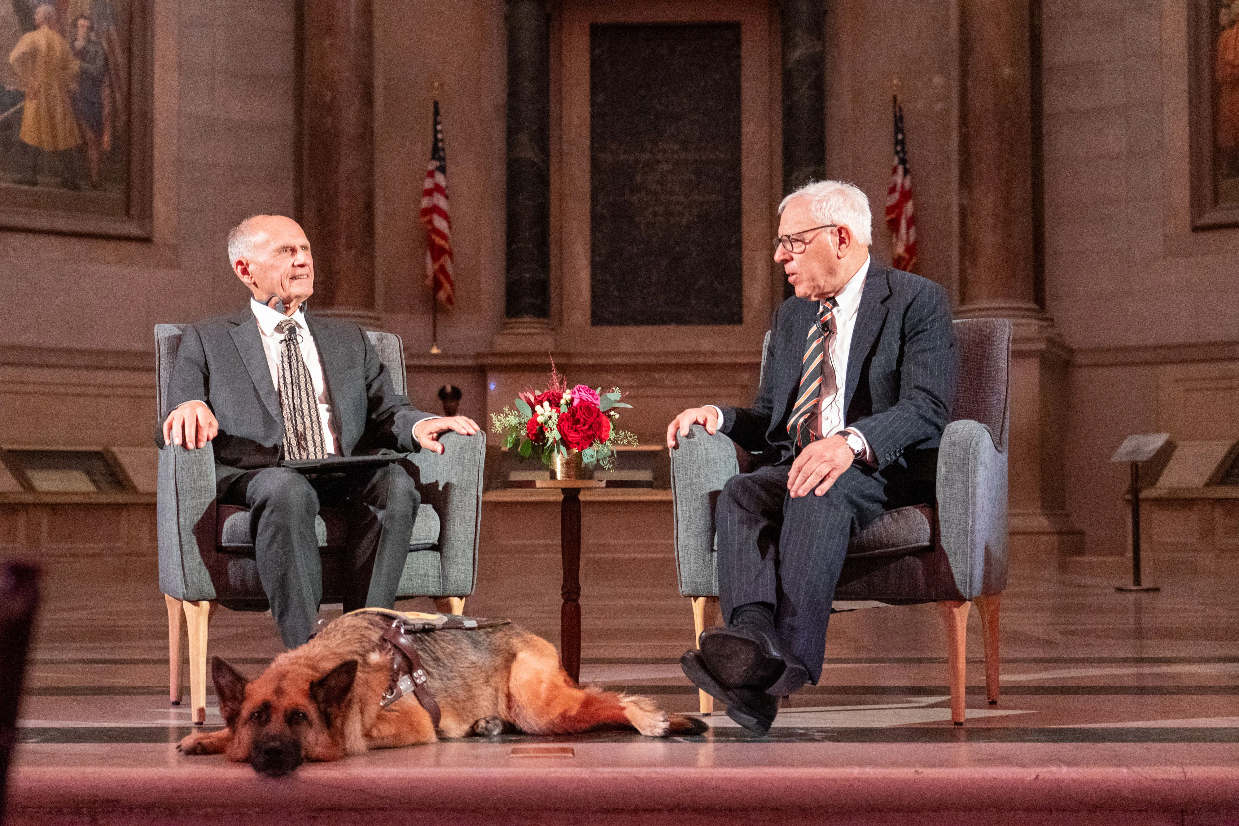 Two people talking across from each other with a table and a dog between them.