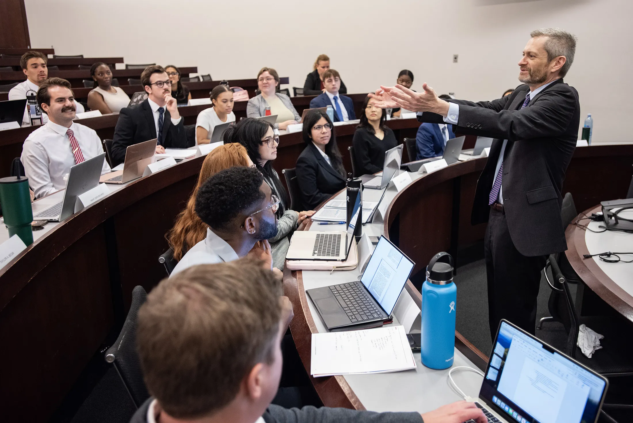 Dean Miles, in a suit and tie, reaches his arms toward students as he teaches in the front of a classroom.