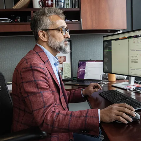 Anup Malani sits at his desk, working from his computer. He wears a maroon plaid sports jacket and blue open-collared shirt.