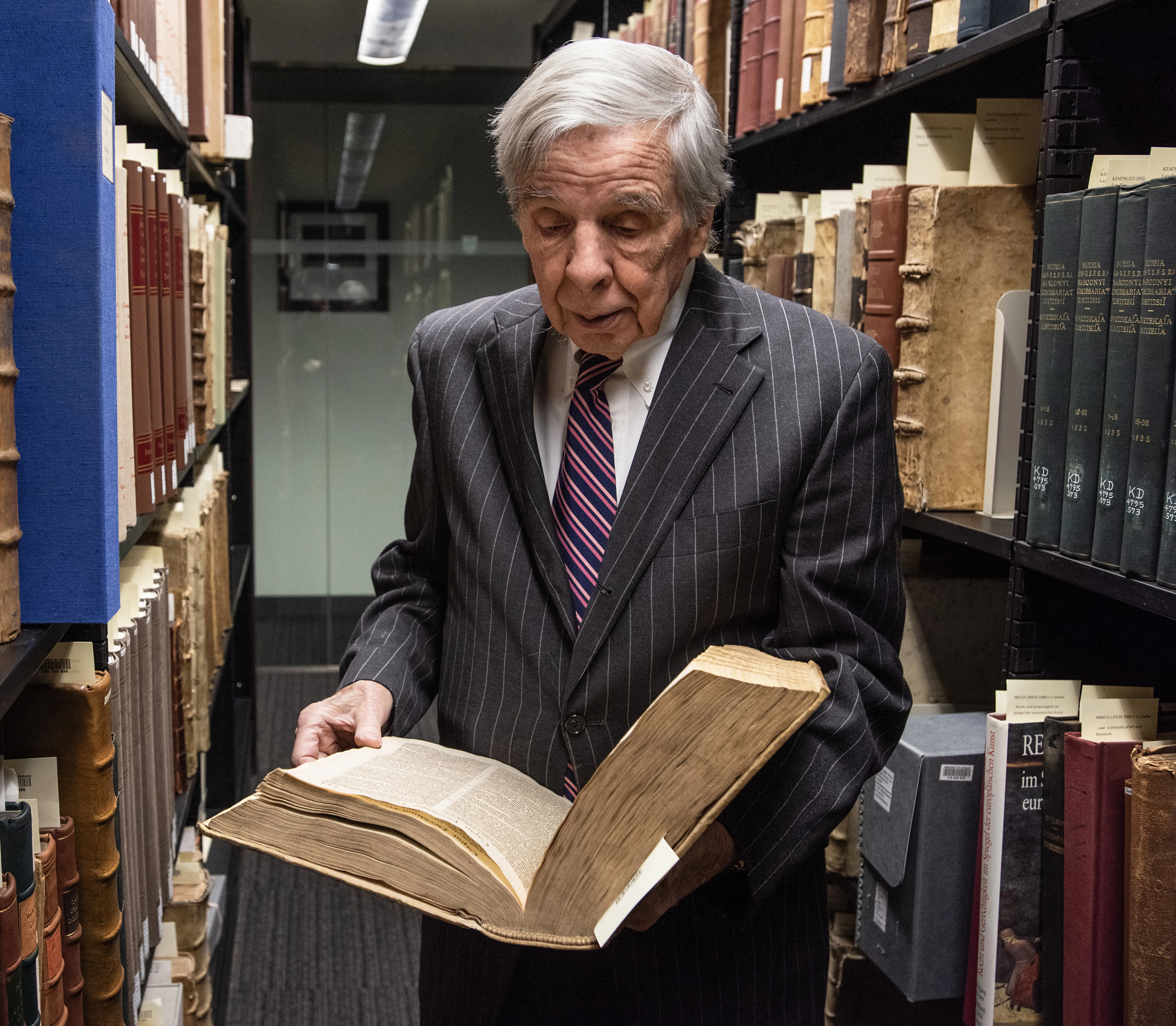 Professor Helmholz, in a dark pinstripe suit and tie, holds an old book. Shelves of rare and old books surround him.
