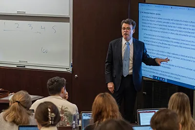 William Birdthistle points to a TV display with text on it. Students look on with their laptops open. Birdthistle wears a blue suit, white collared shirt, and a dotted blue tie.