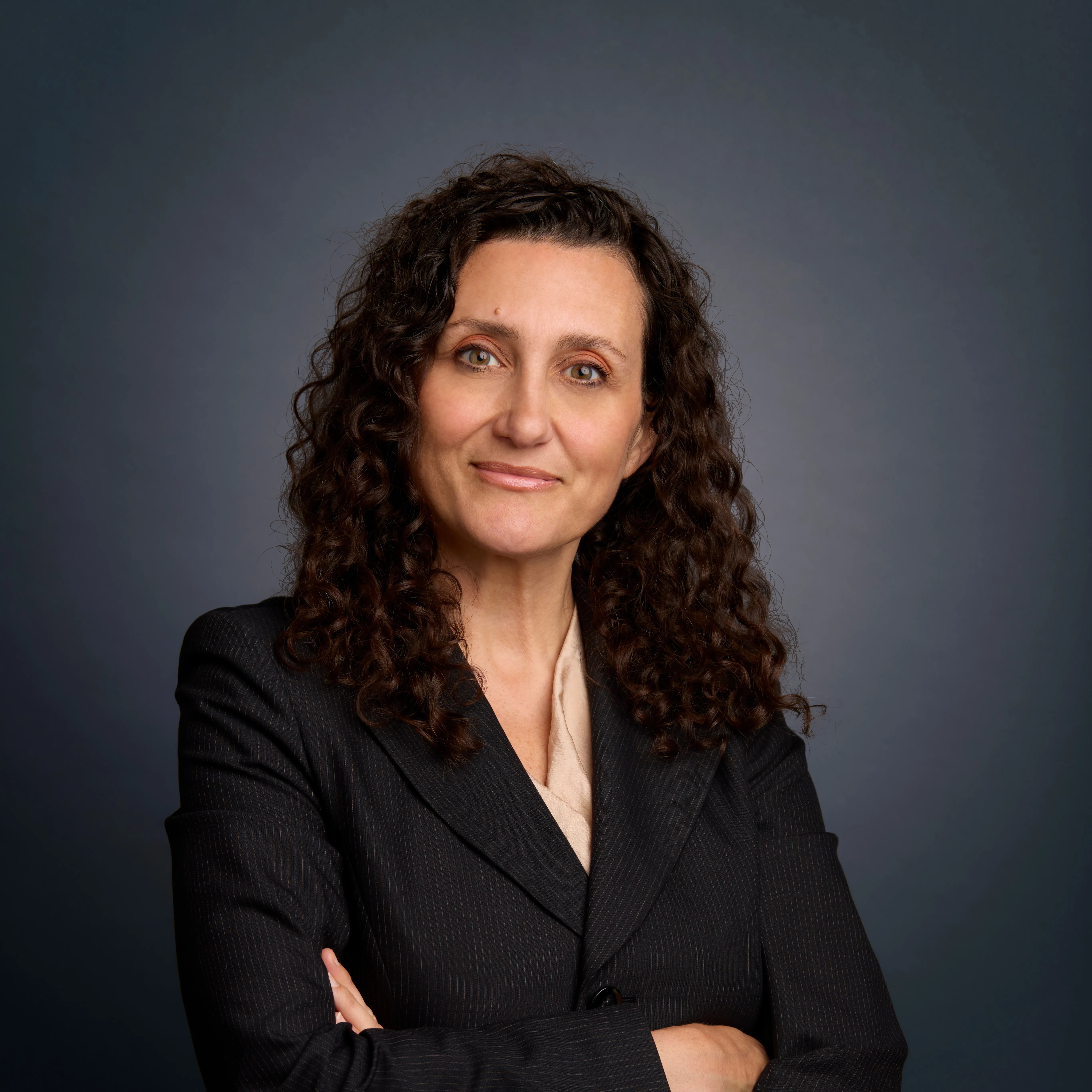 Professional headshot of Sandra Frantzen wearing a dark suit and beige blouse. Her hands are folded across her body.