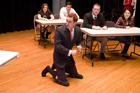 Bill Weaver, in a suit and tie, kneels on top of slippers. Students at desks look on.