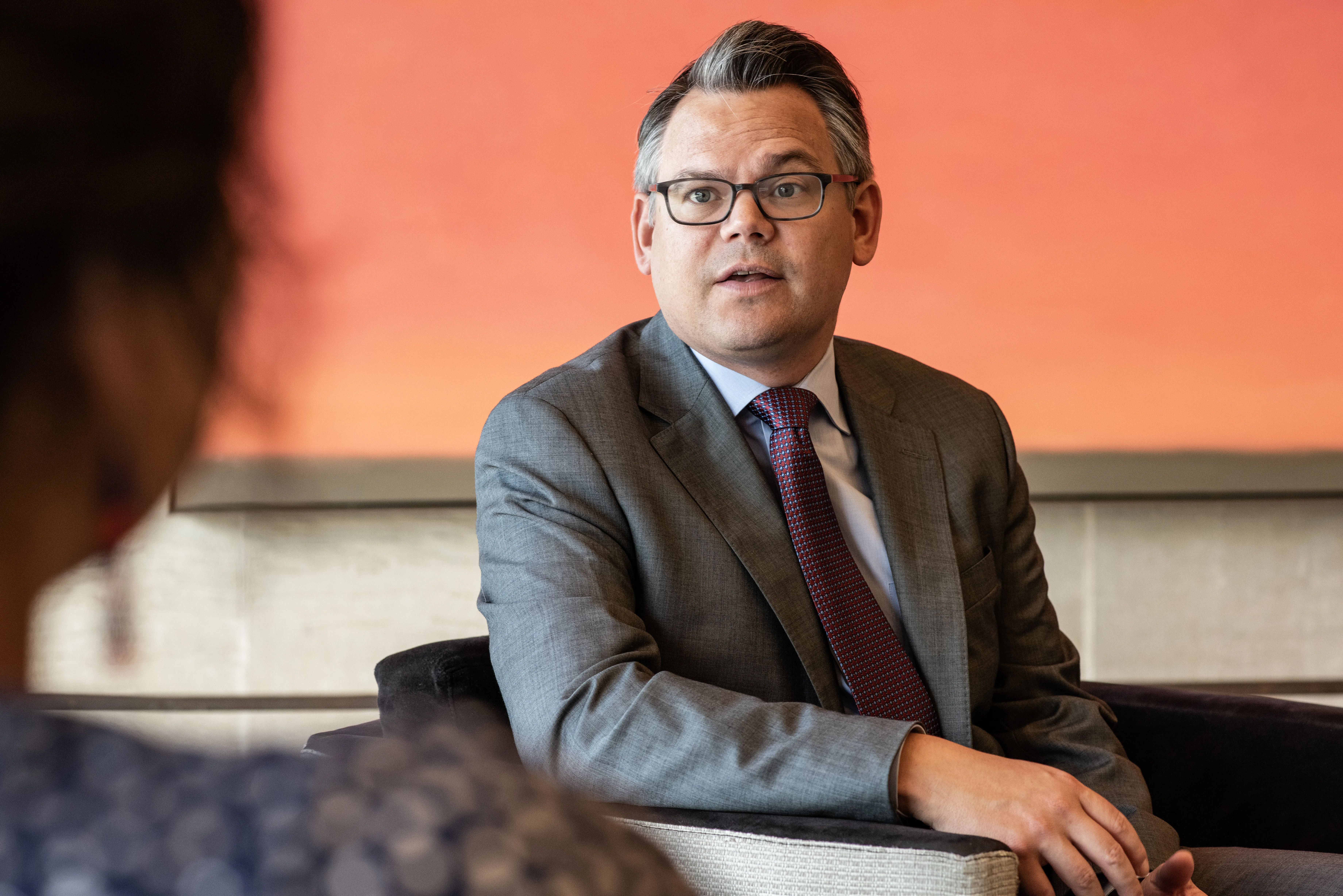 A man wearing a suit and eyeglasses is sitting in a chair in front of a bright orange painting speaking to someone who is in partial view.
