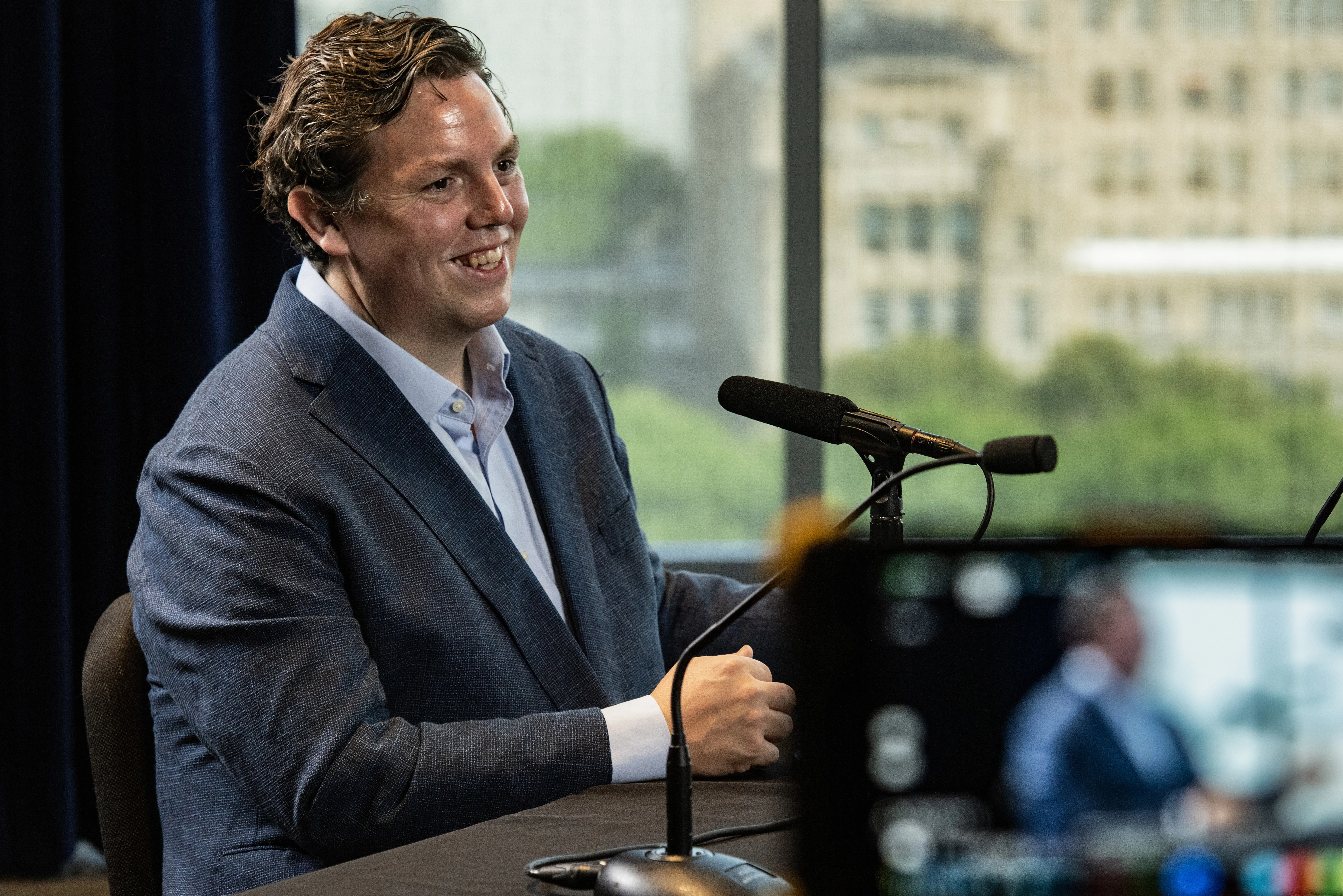 Will Baude wearing a suit is speaking into a microphone while sitting at a desk