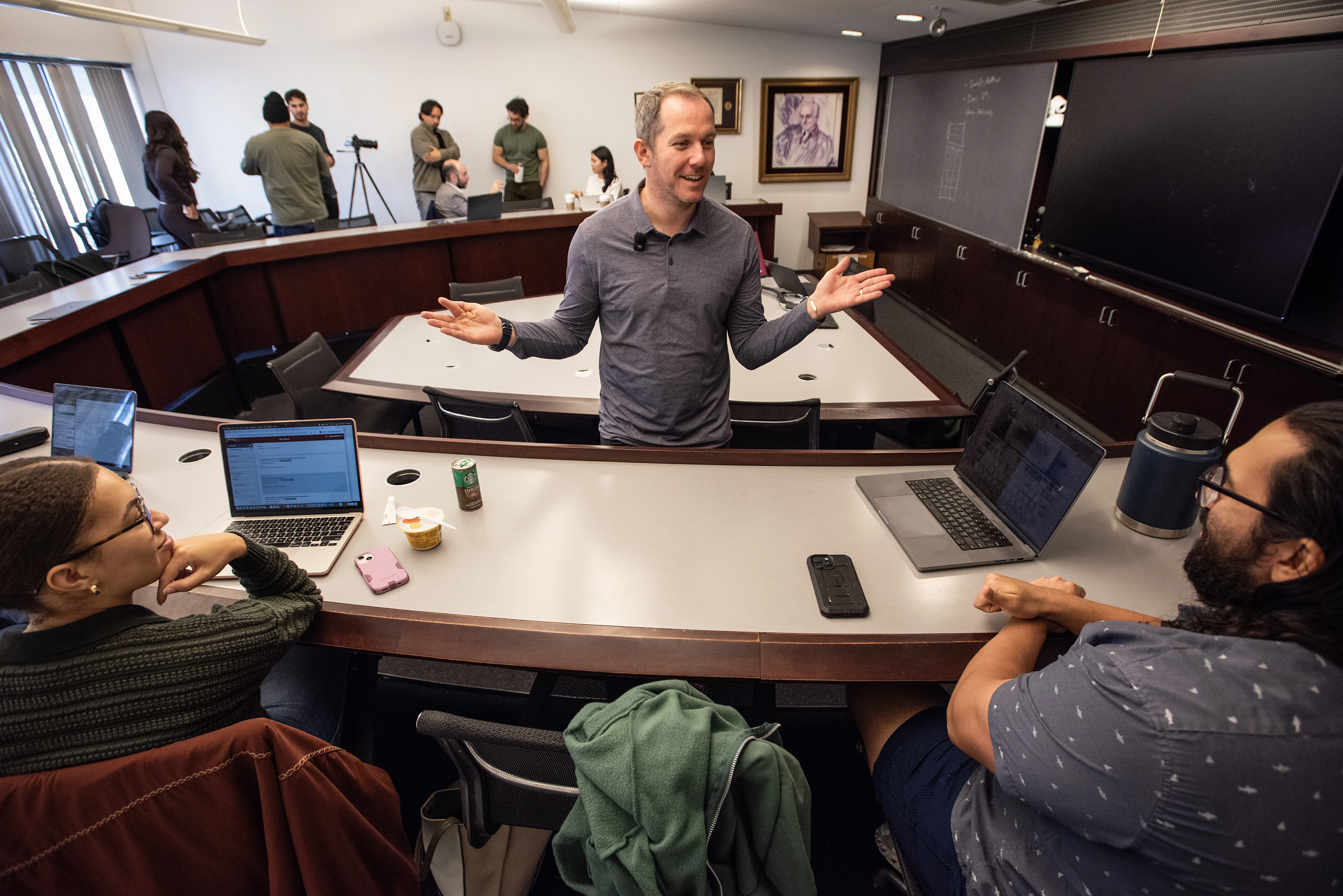 A man in a grey polo shirt addresses a room of students.